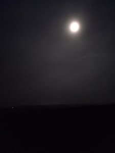 A very grey photograph. There is a line between the dark ground and slightly lighter sky, which fulls the top 2/3 of the photo. The full moon is in the top right of the picture. There's a small white dot on the left hand horizon; I think it's a light in Wrexham.
